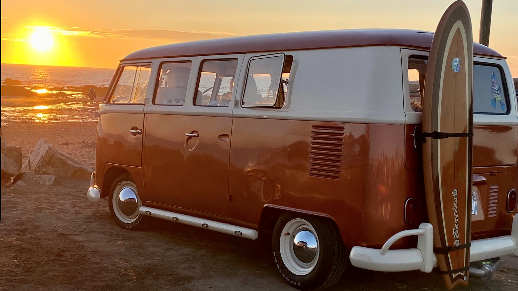 Classic vw campervan in Orange and white with surf board attached upright in the back of the vehicle with a sunset in the background