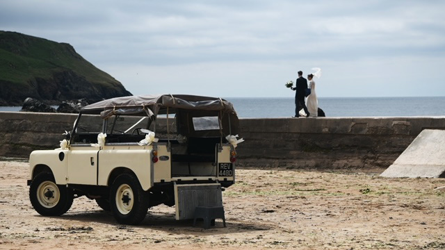 rear view of Classic Land Rover on Devon;s beach. Wedding couple can be seen in the far background walking