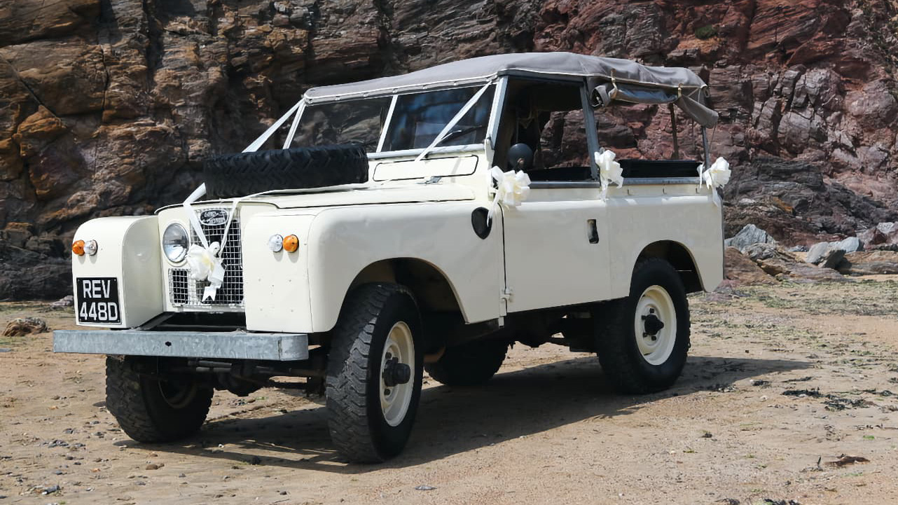 Classic Land Rover in Old English White with white ribbons and spare wheel on front bonnet parked on Devon's beach