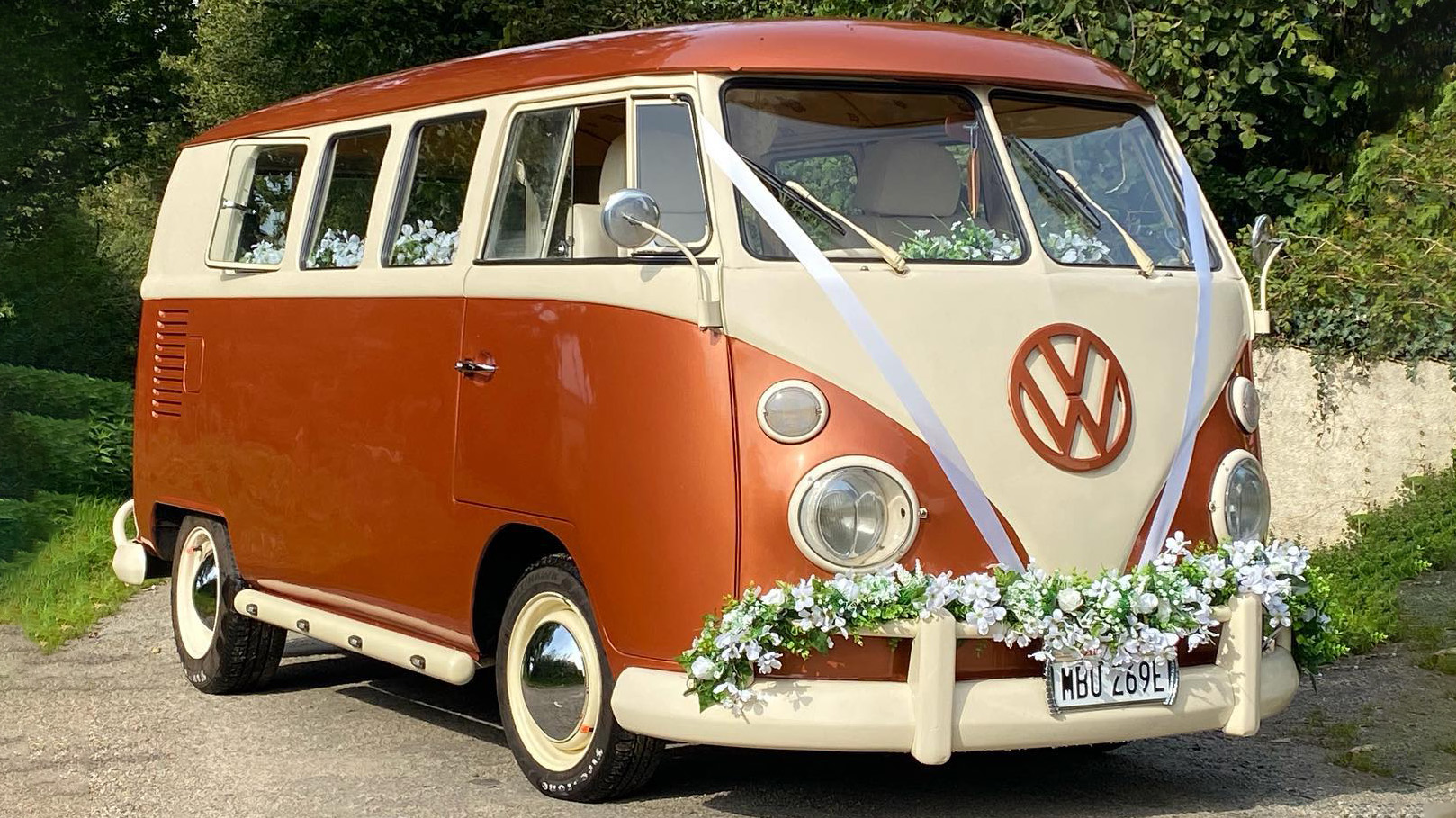 Front right view of classic VW Splitscreen campervan in two tone White and burnt orange decorated with green foliage on front bumper and white wedding ribbons