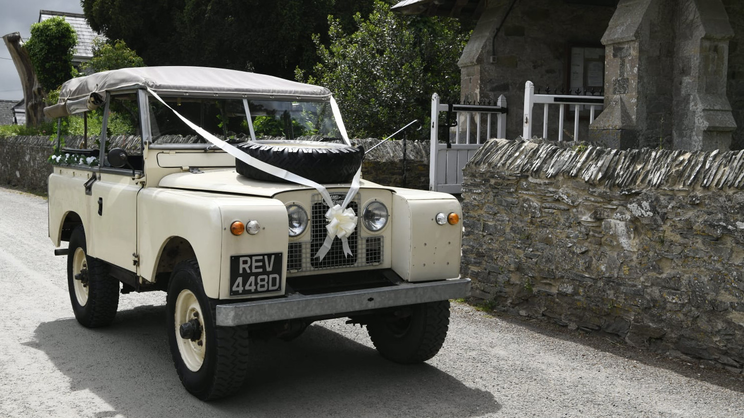 Classic Land Rover in Old English white dressed with white wedidng ribbons parked in front of a church