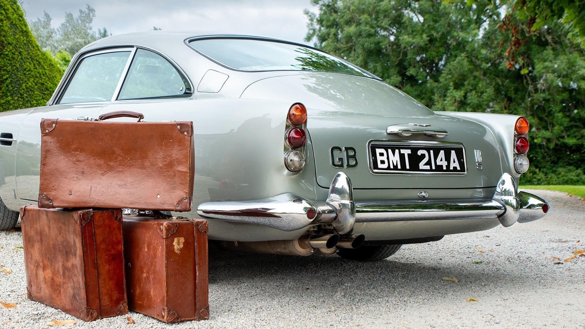 Rear view of classic Aston Martin DB5 in silver with chrome bumpers and three brown vintage suitcases next to the vehicle