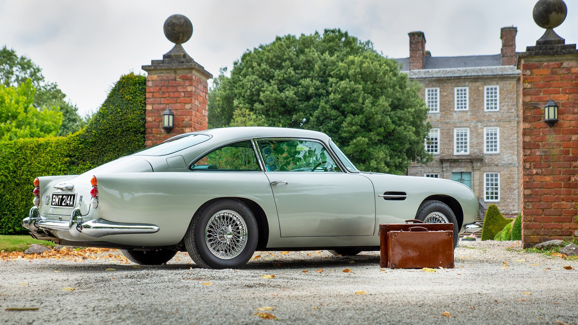 Right side view of a classic Aston Martin DB5 parked in front of a wedding venue with brown vintage cases on the floor.