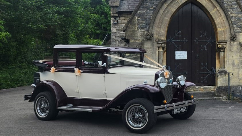 Right side view of a Badsworth in Ivory and Burgundy with black soft top roof open parked in front of a wedding church