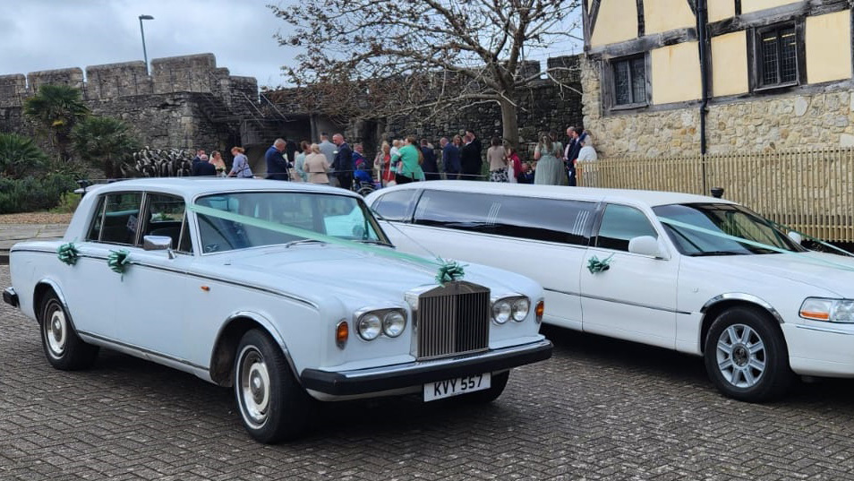 White Rolls-Royce Silver Shadow parked next to a modern White Stretched limousine dressed with matching Green Ribbons