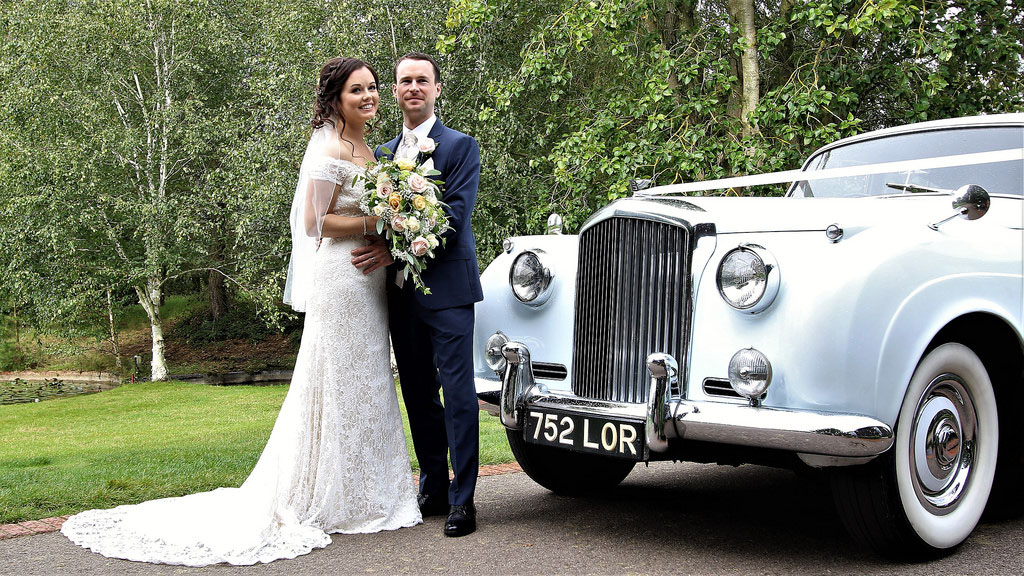 Bride holding a bouquet of flowers with her groom standing in front of a white Bentley S1