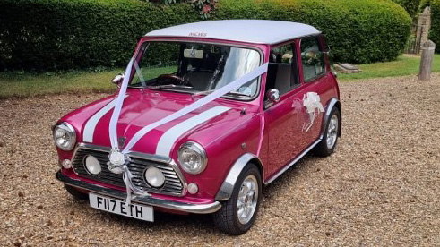 Classic Pink Austin mini wedding car with two white striped on front bonnet and white roof dressed with wedding ribbons and bows attached to door handle.