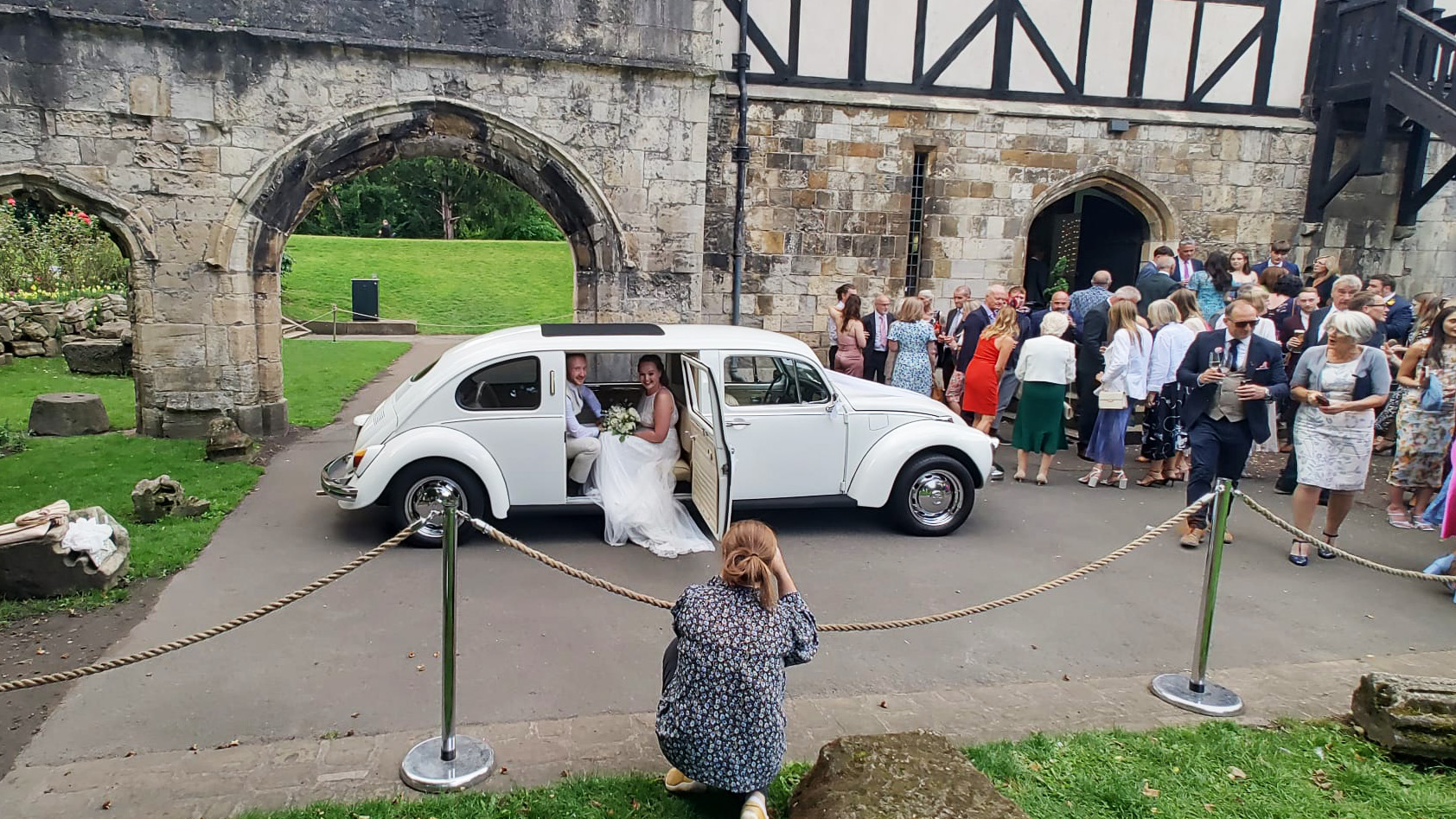 Bride and Groom seating inside a vw stretched limousine with photographer taking photos and wedding guests in the background