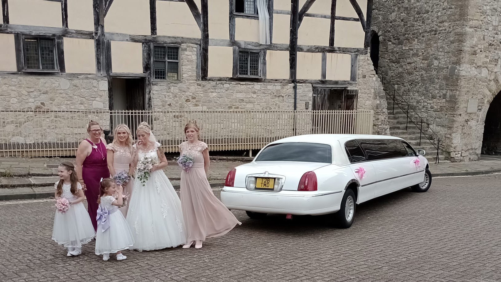 Bride with her bridesmaids and flower girls posing for photos in front of a wedding venue. White stretched limousine in the background.