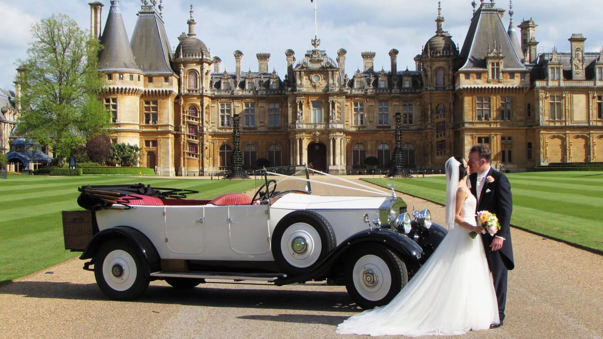 A white vintage Rolls-Royce convertible with Bride and Groom kissing at Waddesdon Manor near Aylesbury shown in the Background