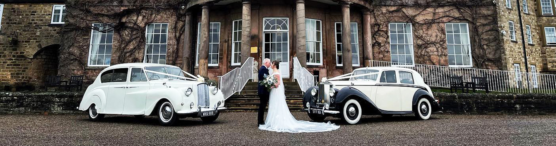 Two wedding cars parked in front of a wedding venue with Bride and Groom holding each others between both vehicles.