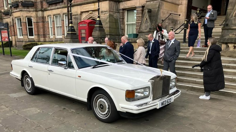 White Rolls-Royce with white wedding ribbons parked in front of registrar in Liverpool with wedding guests in the background