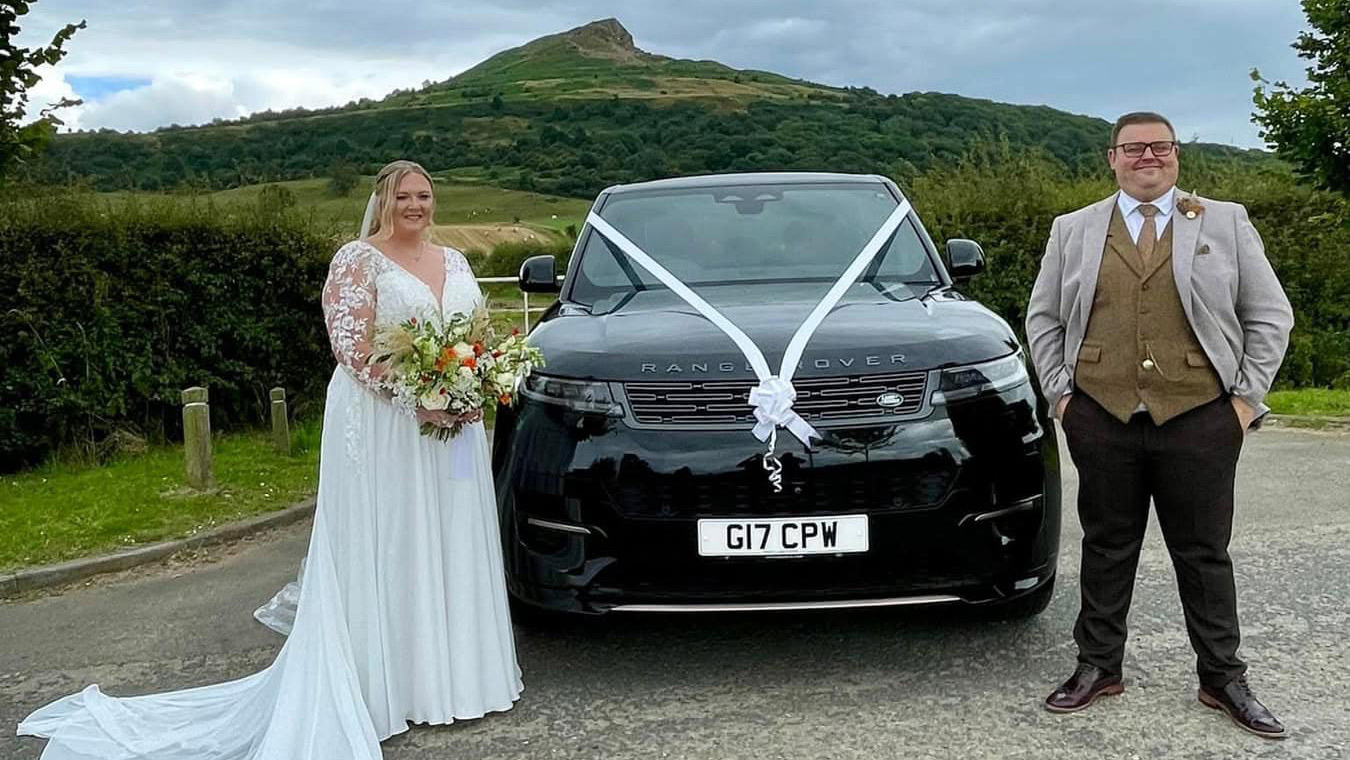 Modern Range Rover in Black with white ribbons and bows with Bride and Groom standing on either side of the vehicle