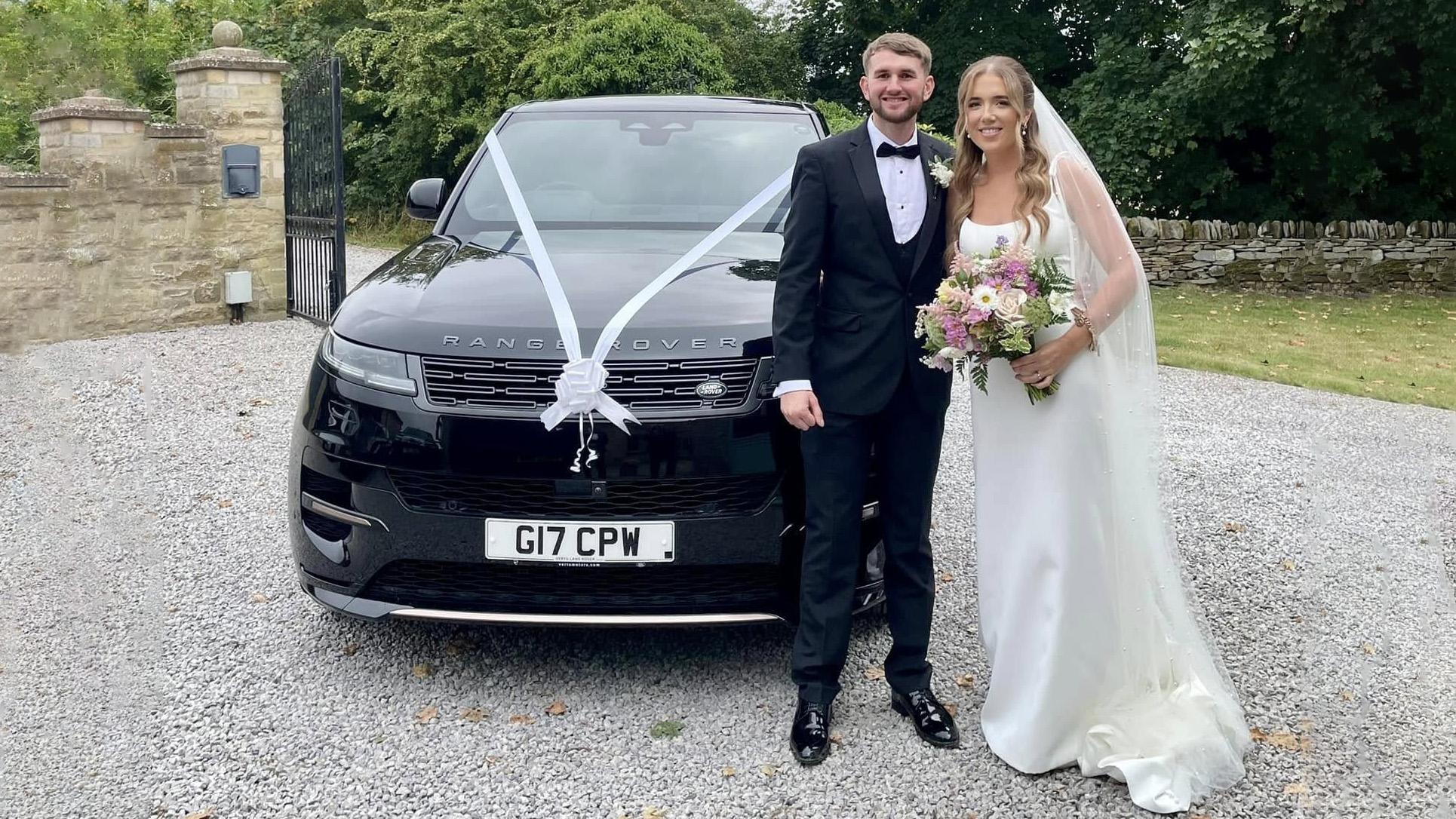 Bride wearing a white wedding dress and holding a bouquet of flowers with the groom in a black suit standing in front of a modern Black Range Rover