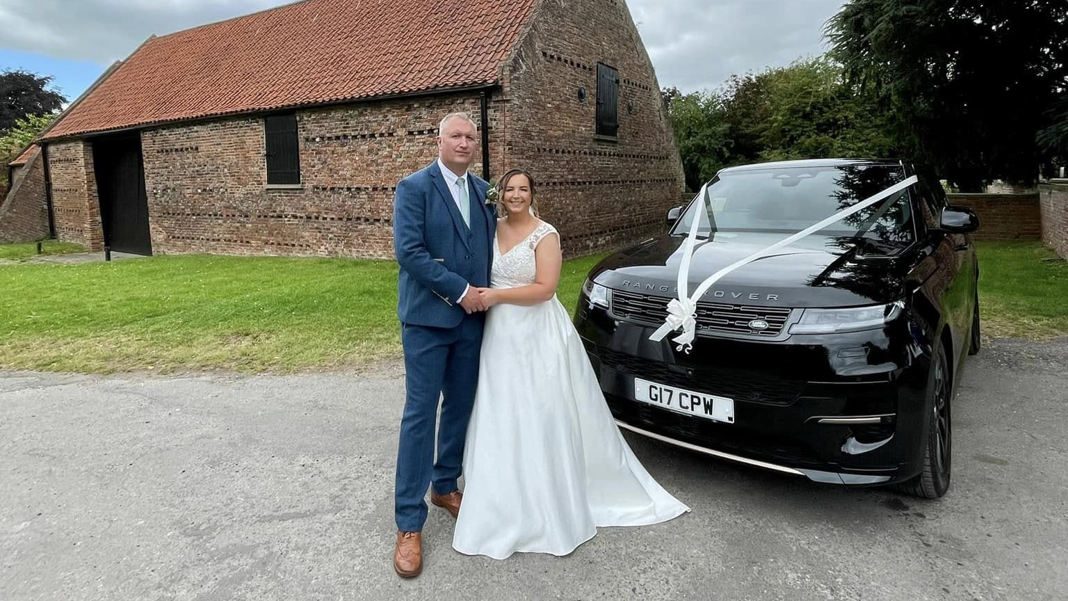 Bride wearing a qhite wedding dress holding her groom wearing a Turquoise blue Suit in front of a modern Black Ranger Rover decorated with white ribbons