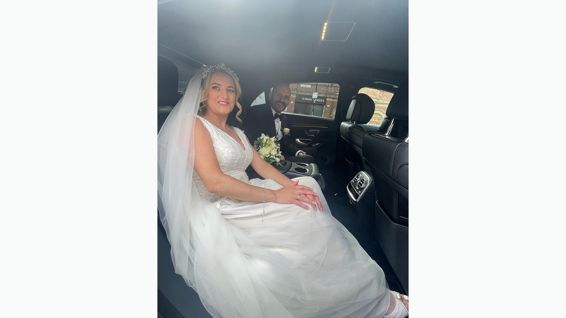 Bride wearing a large white dressed sat in the rear of a Mercedes with her groom