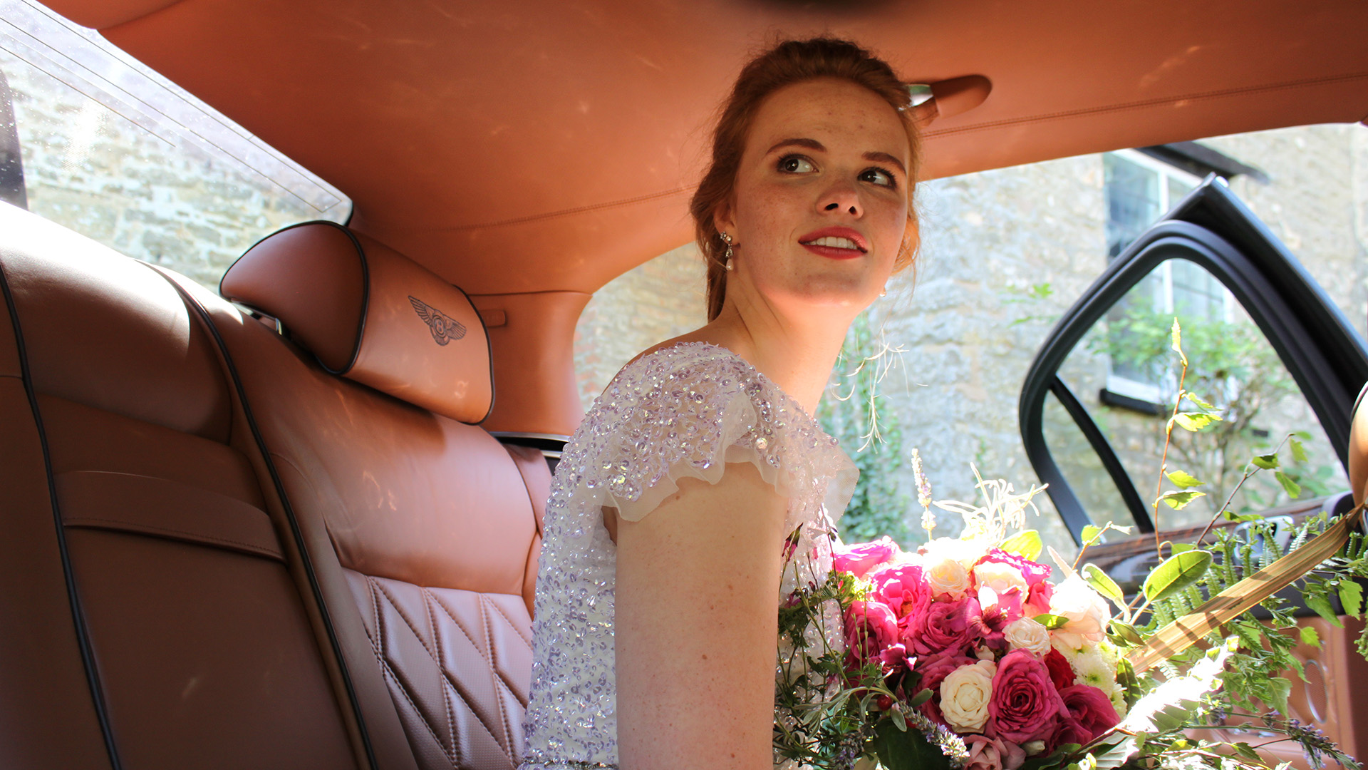 Bride wearing a white dress and holding a colourful bunch of flowers seating inside a Bentley with tan leather interior