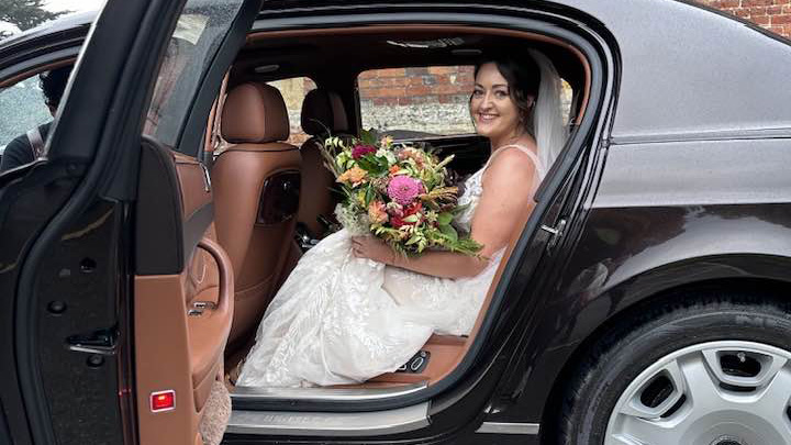 Bride seating inside a Bentley holding a large bunch of flowers with the rear door open