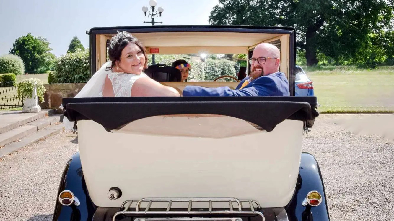 Bride and Groom seating inside a vintage imperial car with roof down looking backward towards the photographer.