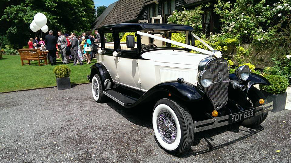 Badsworth Landaulette in Black and Ivory decorated with Ivory ribbons parked in front of a church with wedding guests in the Background