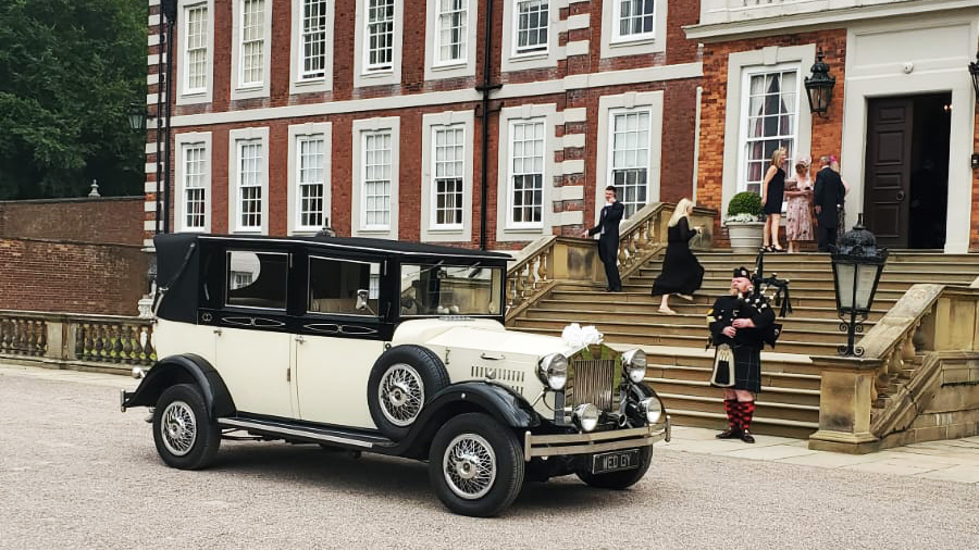 Ivory imperial wedding car with black wheel arches and roof parked in front of a wedding venue in Liverpool. Bagpipe player in the background with some wedding guests