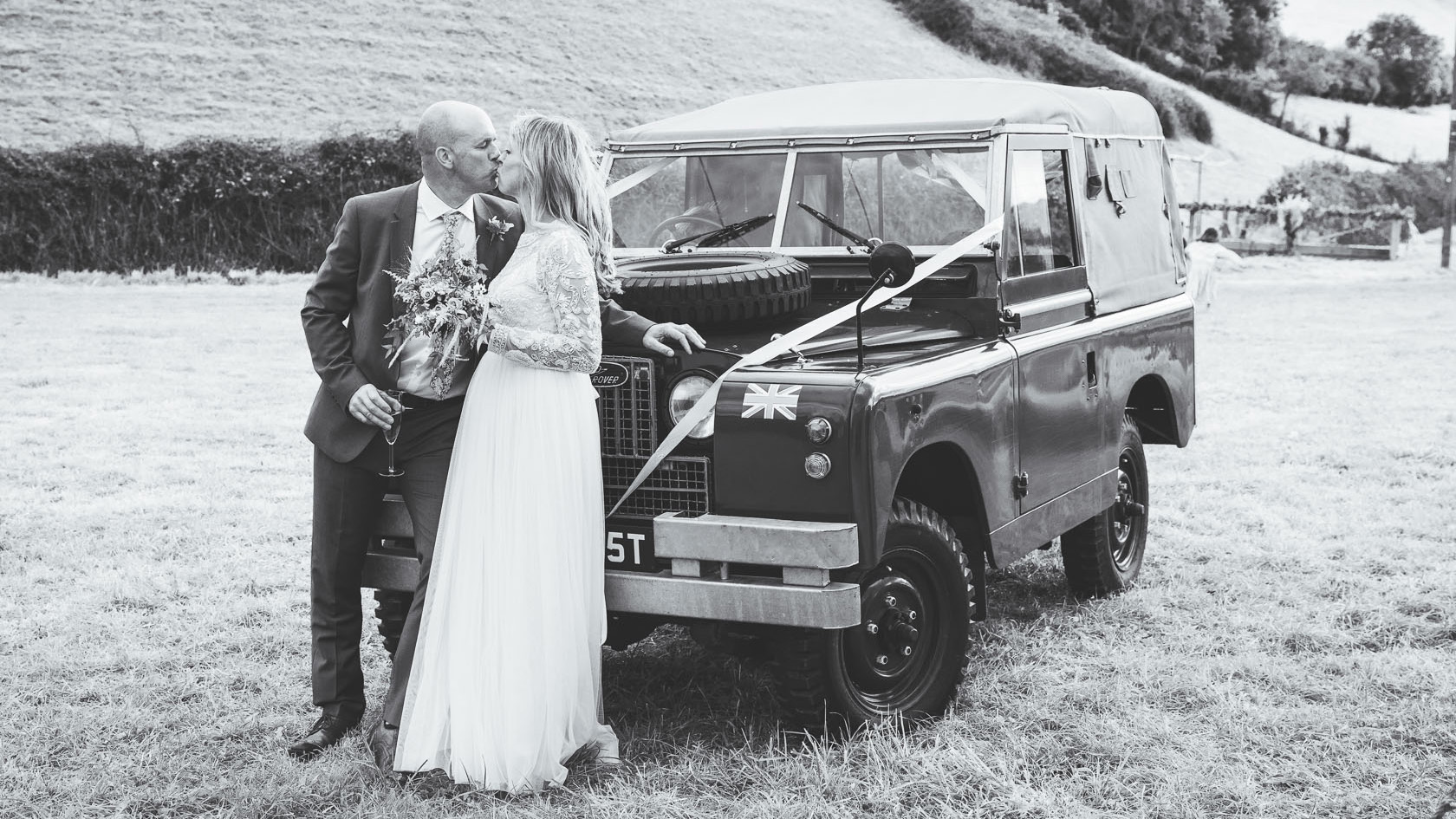 Black and White photo of a Bride and Groom kissing in front of a classic Land Rover dressed with wedding ribbons