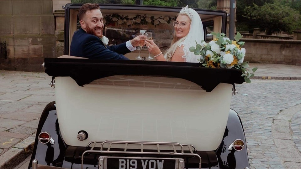 Bride and Groom sat in the back of a convertible vintage car with the roof down holding a glass of Champagne