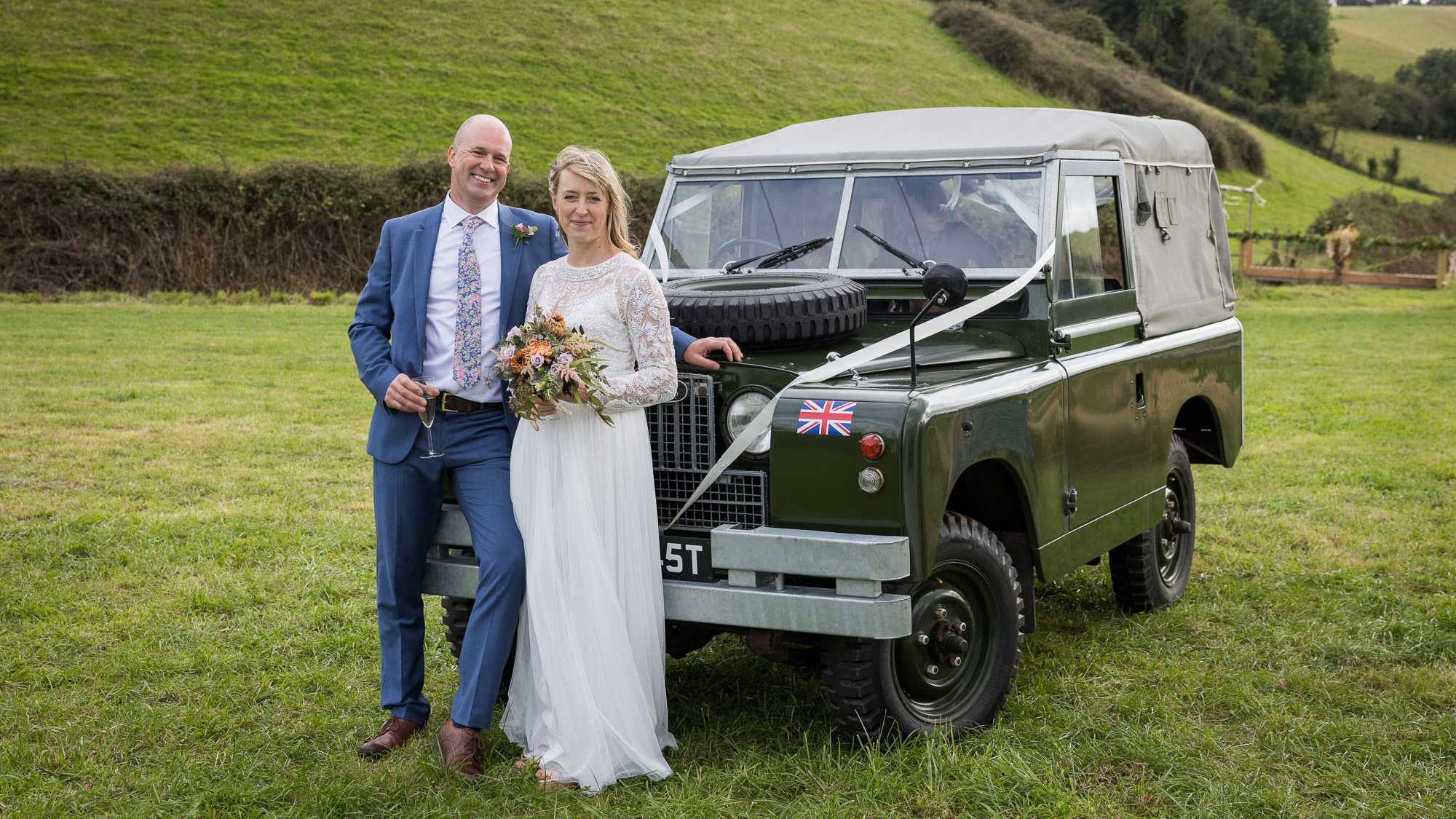 Bride and Groom standing in front of a classic Land Rover in Green decorated with white wedding ribbons