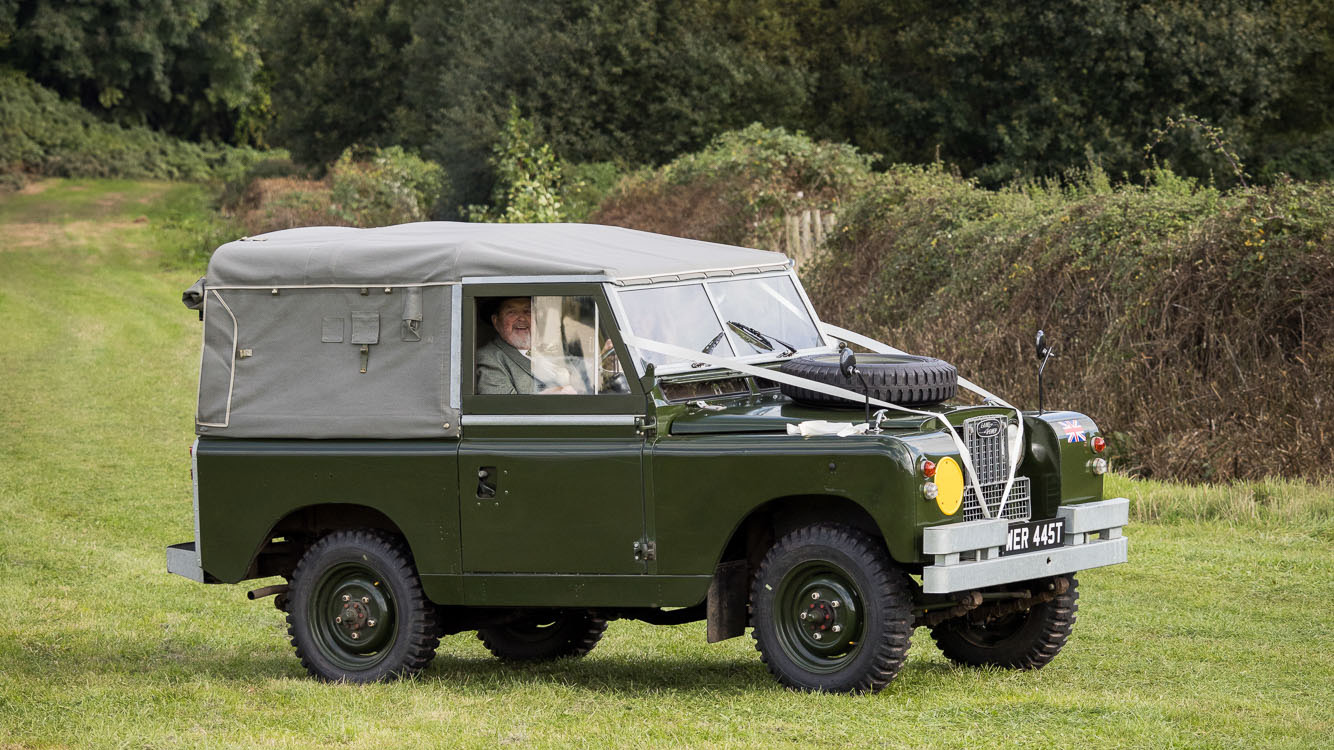 right side view of a green Classic Landrover with a light silver canvas and spare wheel mounted on bonnet.