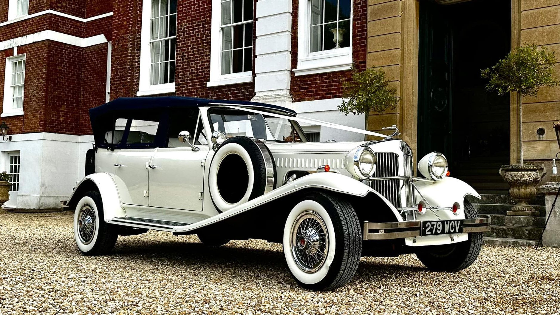 White vintage convertible Beauford wedding car decorated with white ribbons parked in front of Pynes House in Exeter. White wall tires, spare wheel mounted on the side.