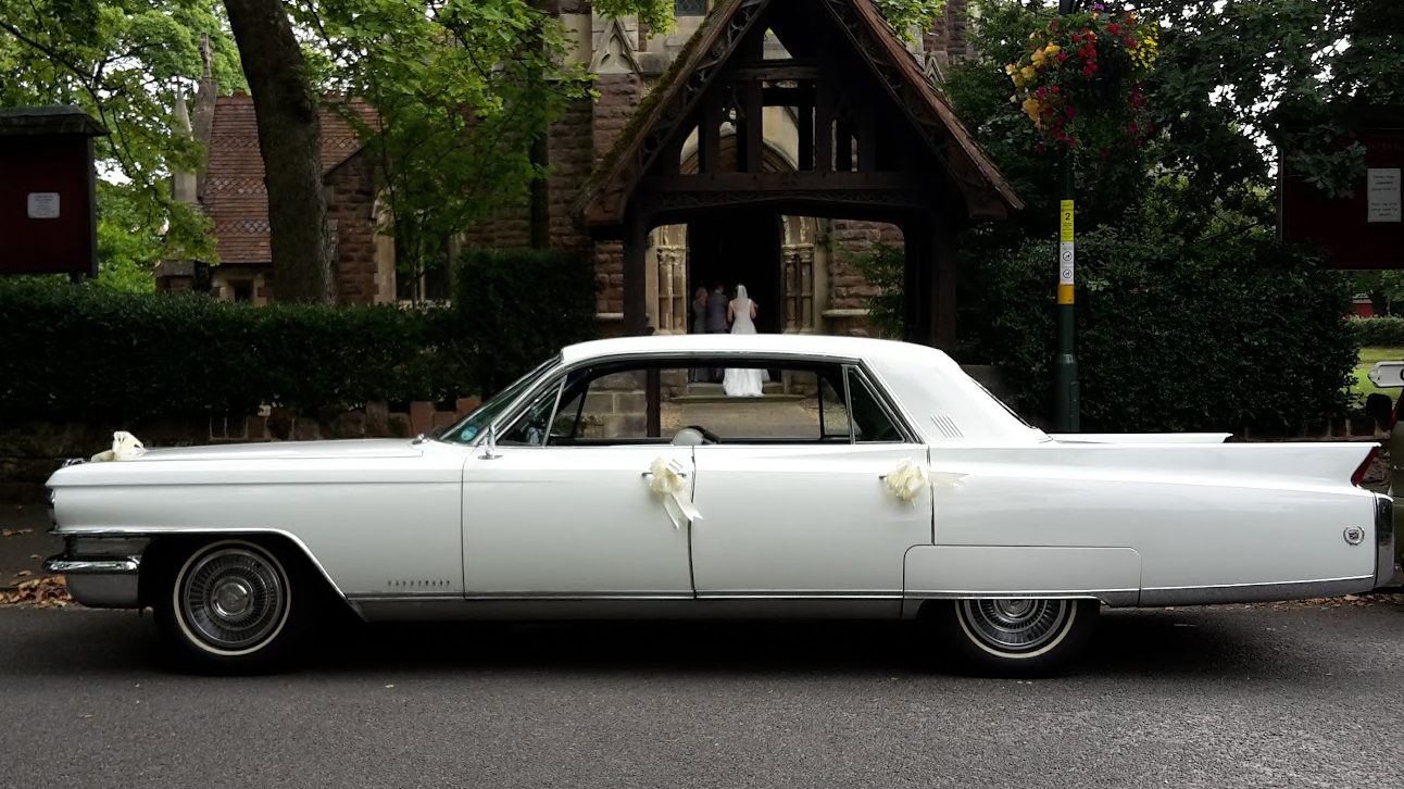 Left side view of a classic White American Cadillac Fleetwood parked in front of a church with Bride walking in the church in the far background