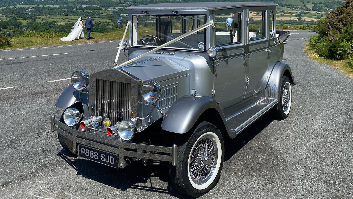 Vintage Imperial wedding car dressed with white ribbons parked on the side of the road with Bride and Groom in the very far background