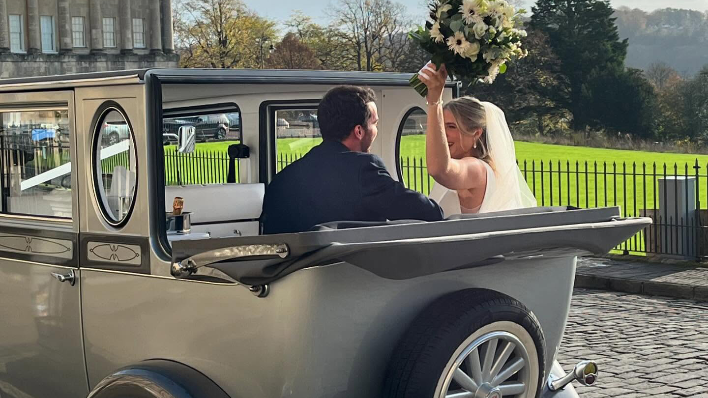 Bride and Groom seating inside a vintage style imperial with convertible roof down and bride holding a bouquet of flower in the air.