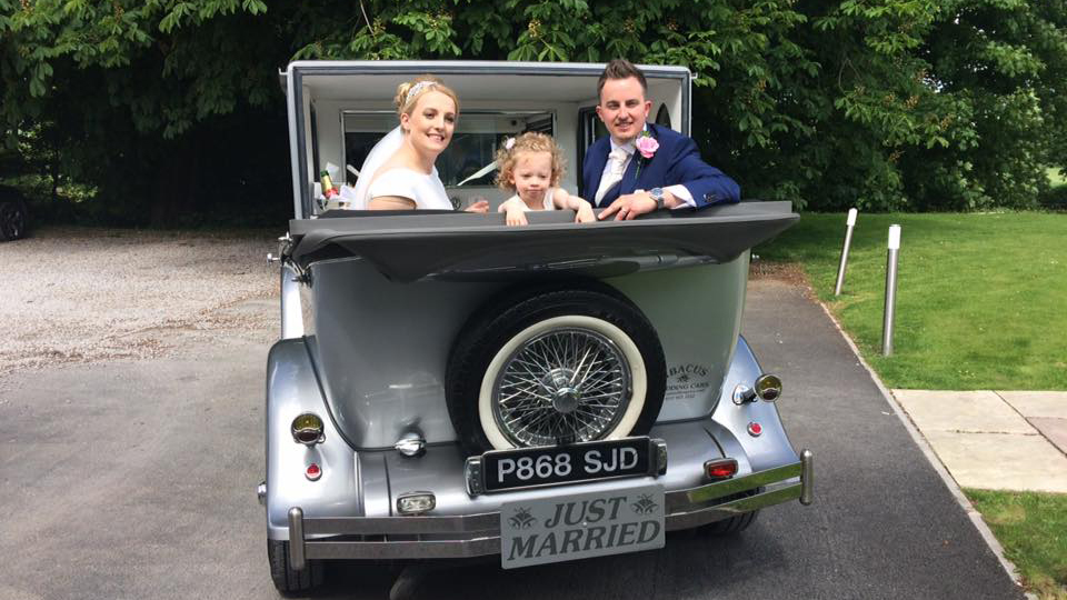 Bride and Groom with their daughter seating inside an Imperial wedding car looking backward with the convertible roof down