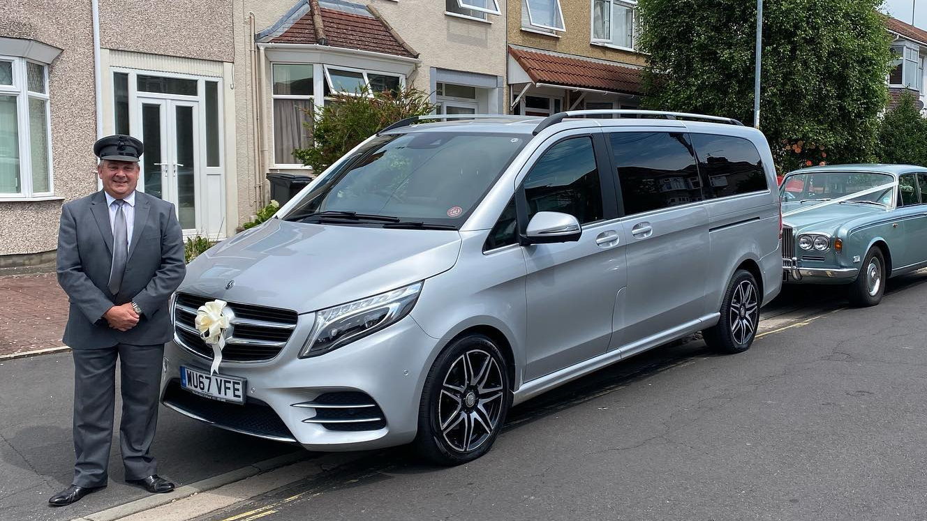 Modern Silver Mercedes V-Class dressed with Ivory ribbons and fully suited chauffeur standing in front of the vehicle