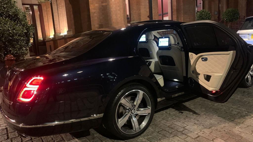 Modern Bentley Mulsanne at dusck with rear lights on and passenger door open showing cream door card and illuminated TV