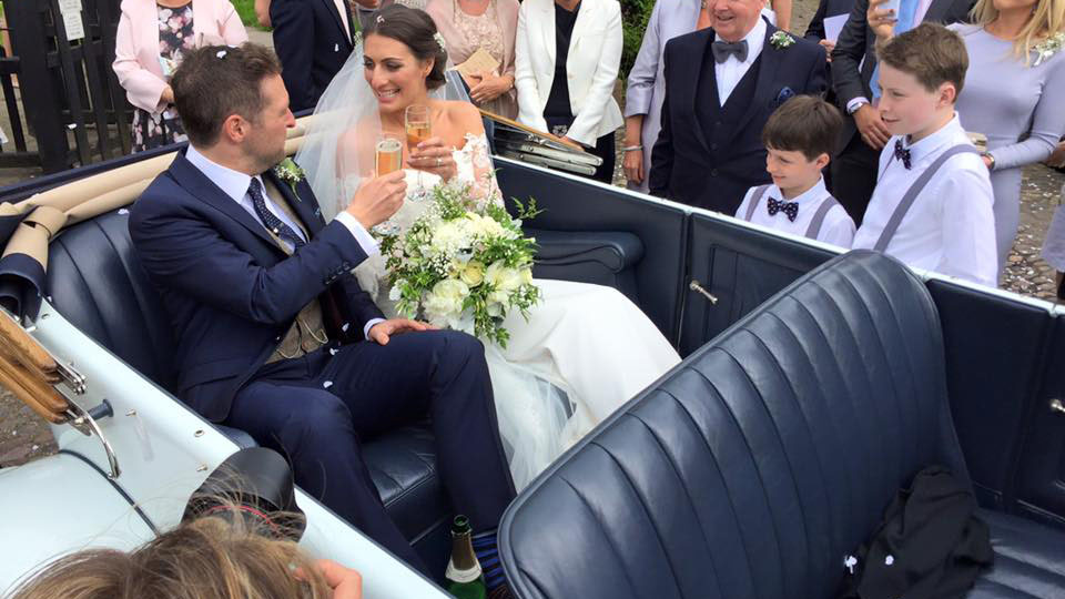 Bride and Groom seating inside vintage Rolls-Royce with roof down cheering and smiling with wedding guests all around the vehicle.