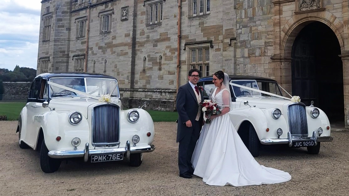 Two identical Classic 7-seater Austin Princess Limousines in Black and White dressed in matching ivory ribbons and bows. Both vehicles are parked in front of a wedding venue with Bride and Groom standing in the middle of the cars.