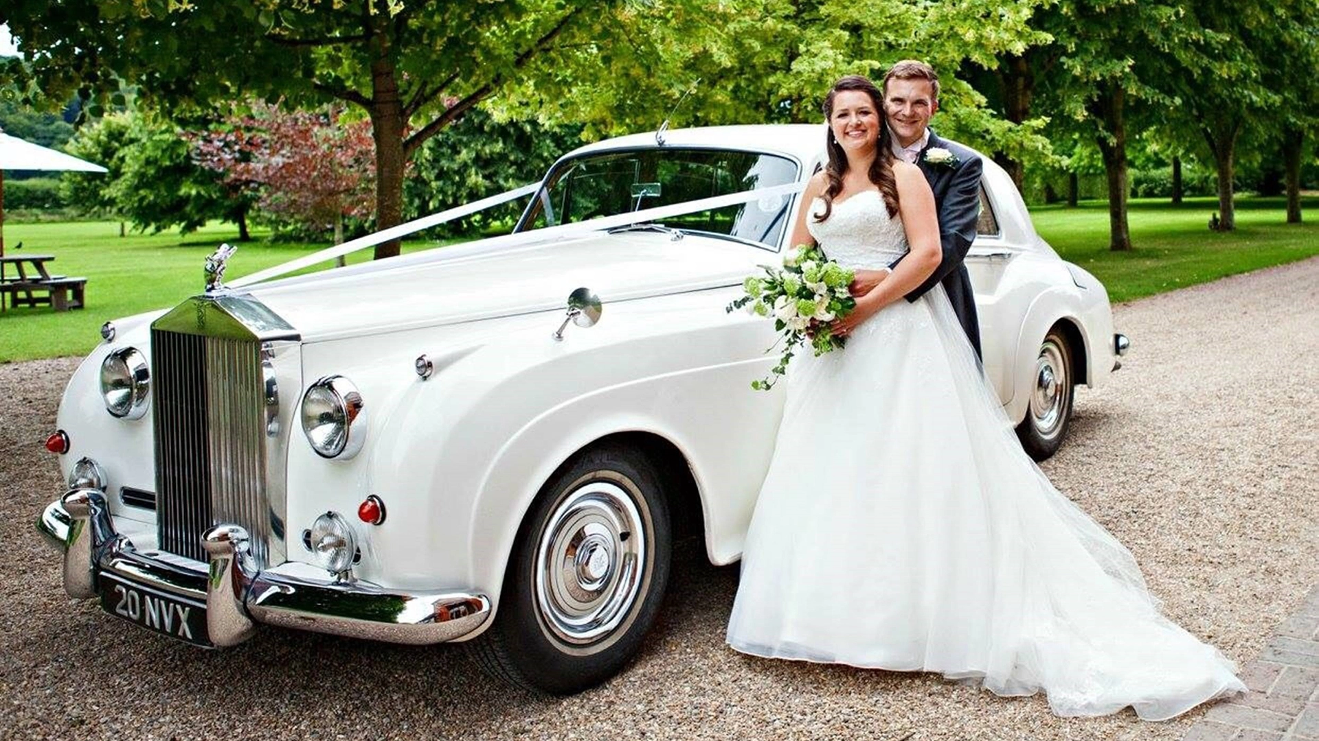 A White Classis Rolls-Royce Silver Cloud in white with a large and imposing chrome front radiator grille and chrome front bumper. The car is dressed in traditional white ribbons. Bride and Groom are holding each other’s smiling.