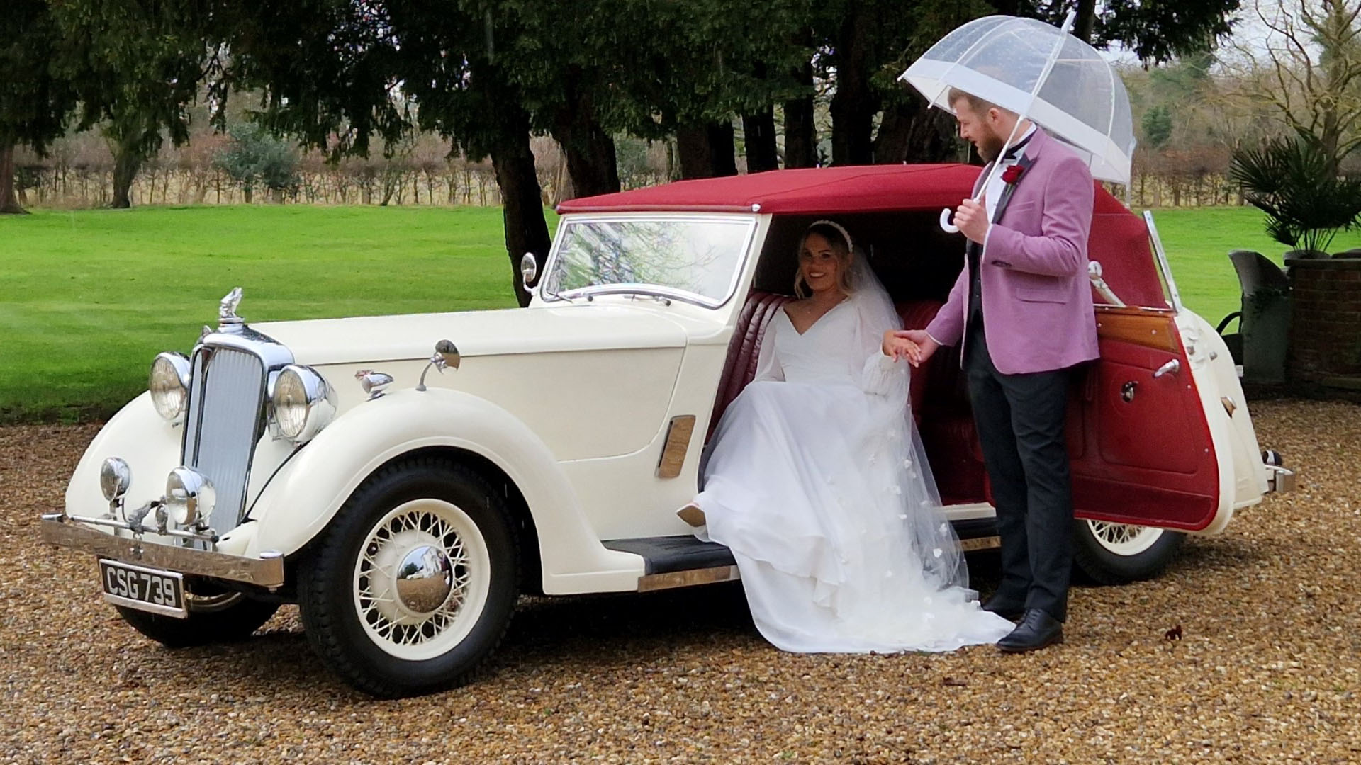 Bride wearing a white dress seating inside a vintage Rover with groom holding an  umbrella in one hand and help his bride with the other