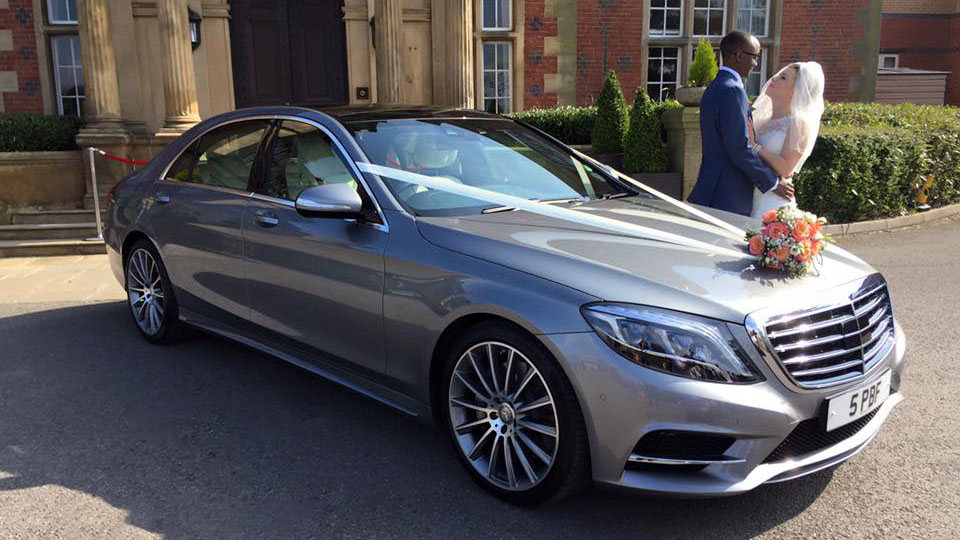 Bride and Groom holding each others behind a silver Mercedes. Car is decorated with Ivory ribbons and bridal bouquet on bonnet.