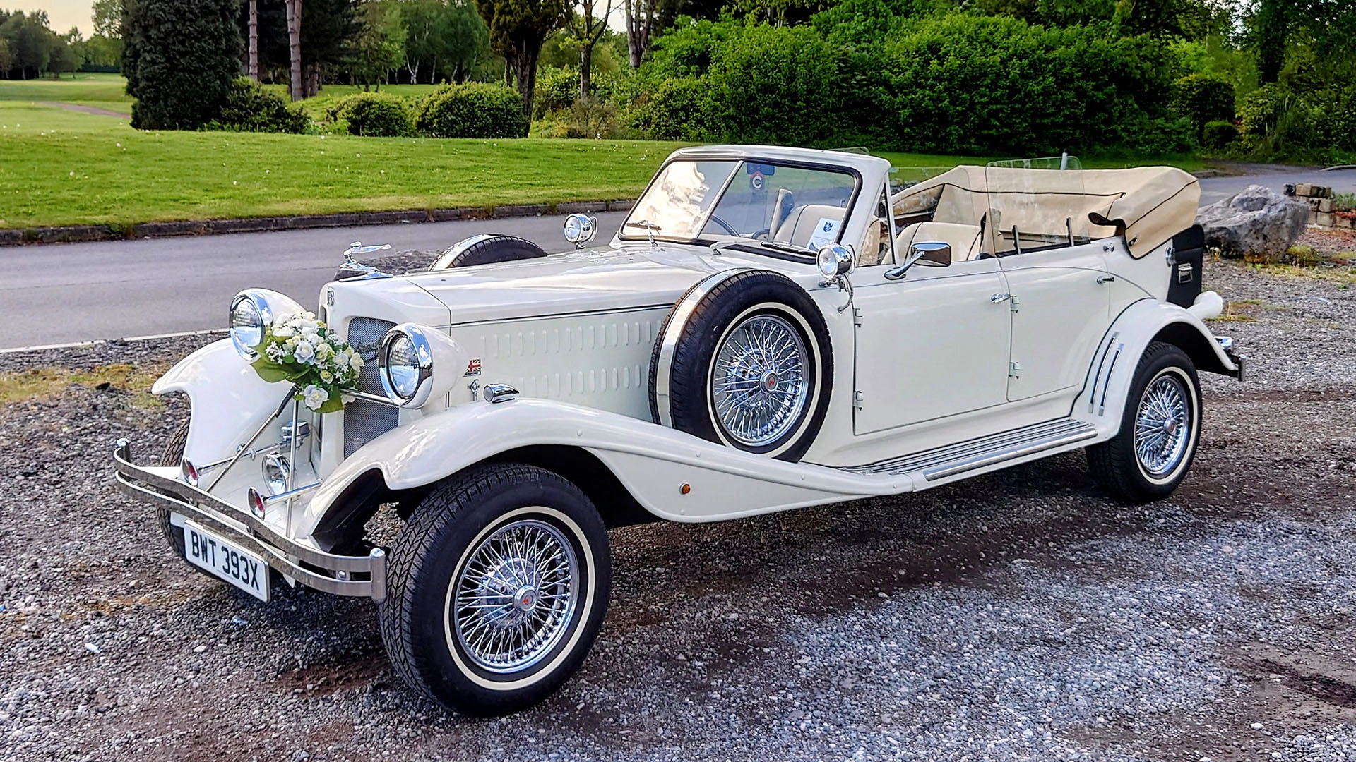 Cream Vintage-style Beauford with a fully-convertible roof. Spare wheel is mounted on the side of the vehicle. Vehicle is decorated with wedding flowers attached to the front grille, white wall tires and cream leather interior.