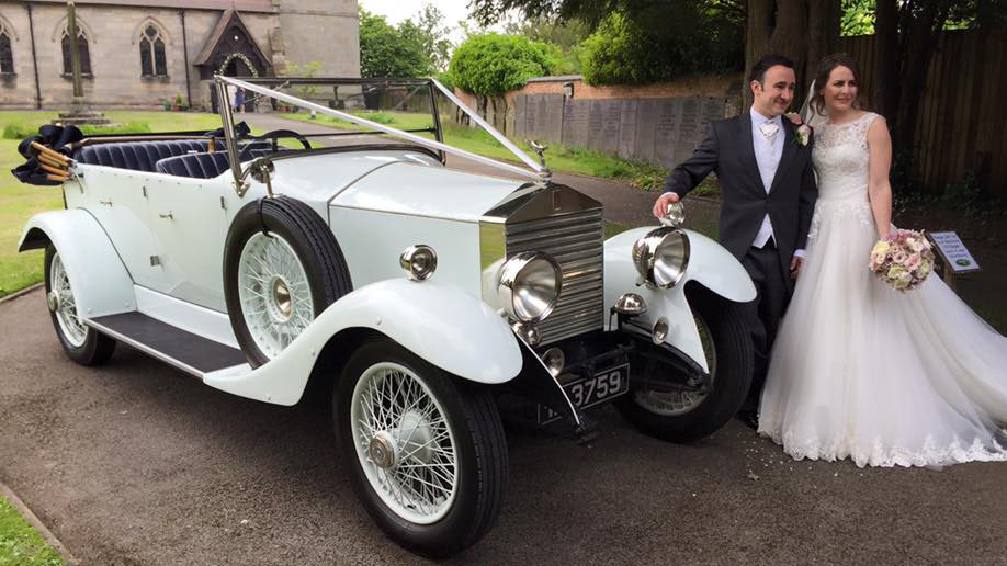 Bride and Groom standing next to a White vintage Rolls-Royce convertible. Church can be seen in the far background