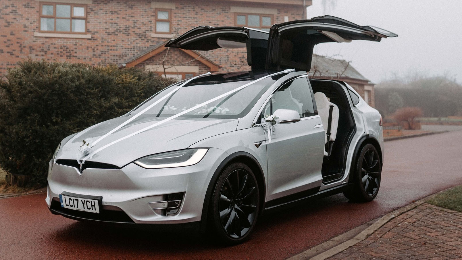 Left side view of a Silver Tesla X decorated with white ribbons and Gull Wings open