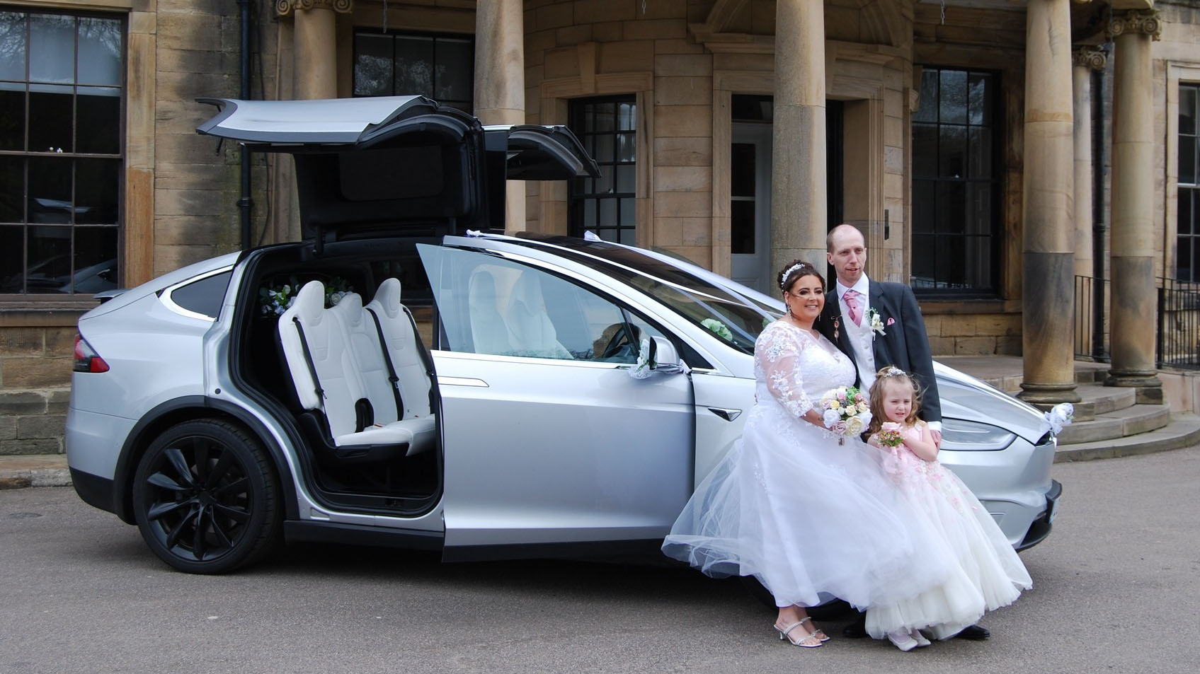 Right side view of a silver Tesla X with Gull wings open and Bride and Groom standing by the vehicle