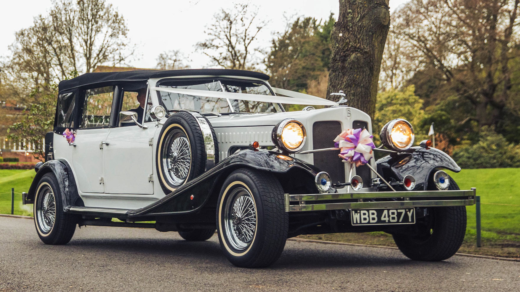 vintage Beauford in white with black wheel arches and black soft top roof decorated with wedding ribbons and head lights switched on.