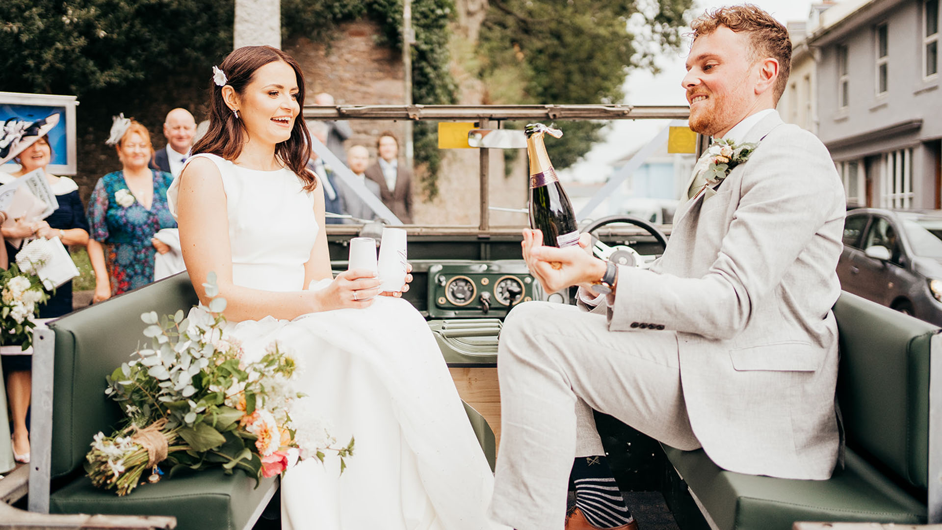 Bride and Groom inside classic Land Rover with groom opening a bottle of champagne