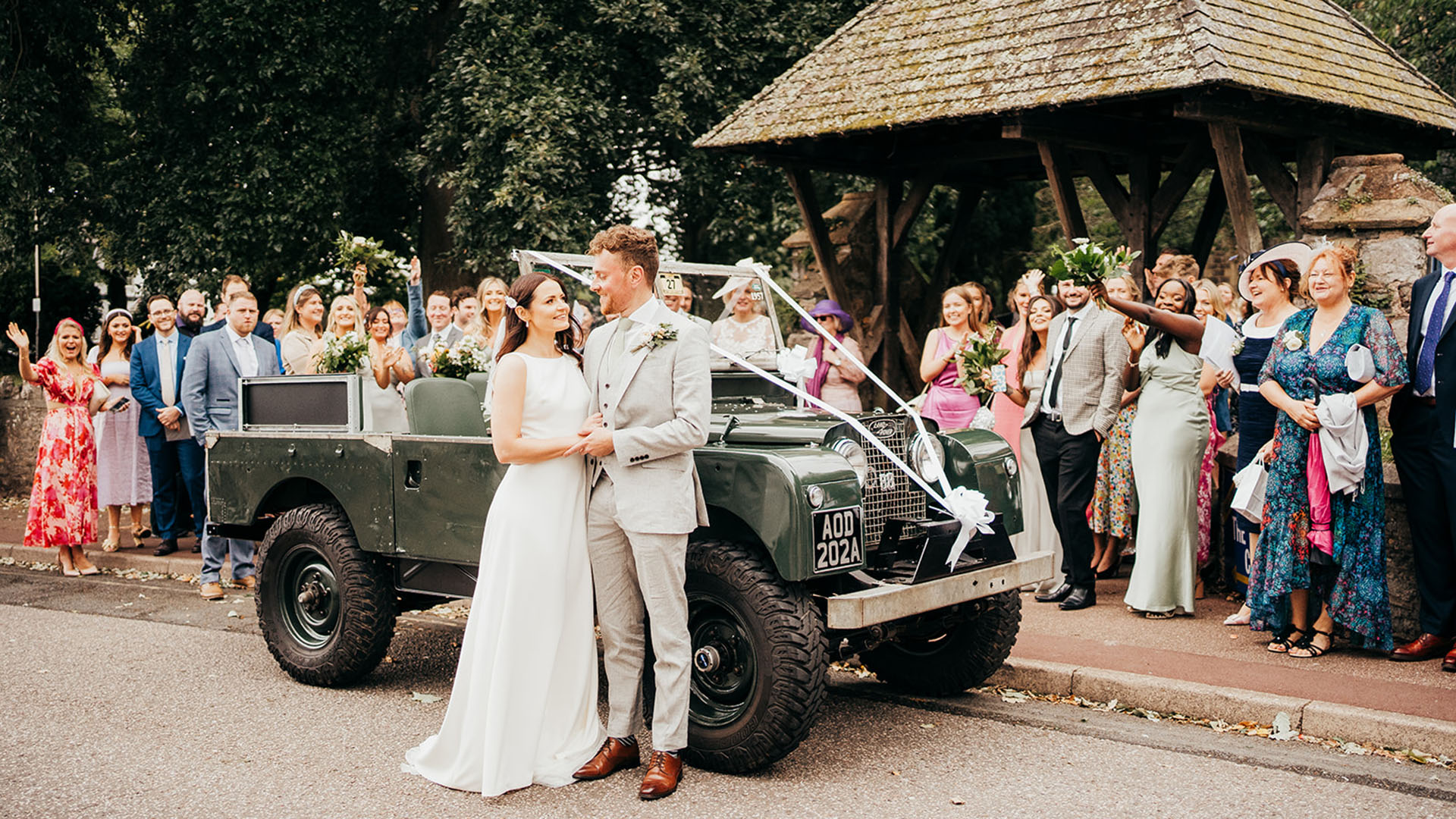 Bride and groom standing in front of an army green Land Rover with wedding guests in the background