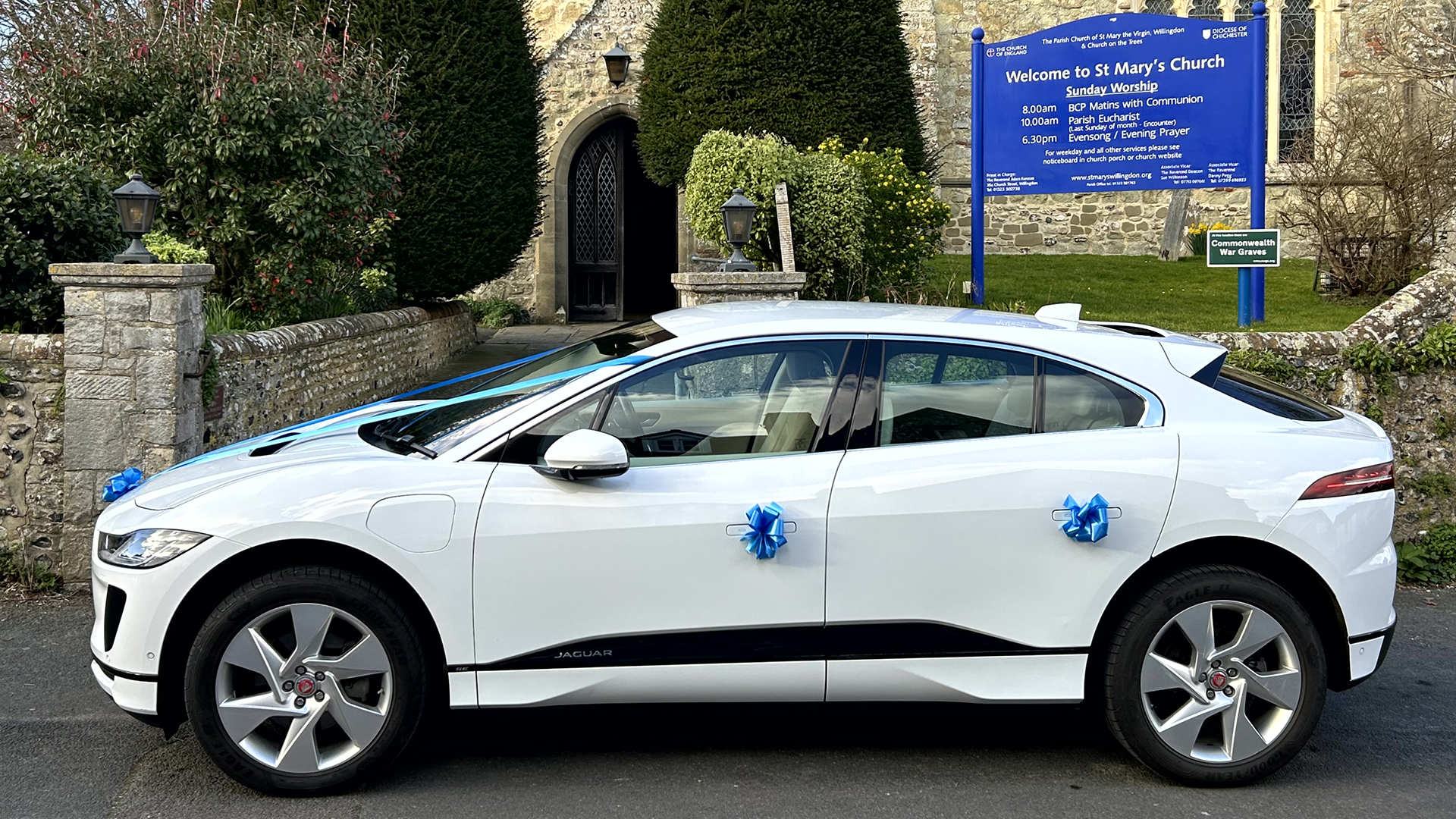 Right side view of a white Jaguar I-Pace with blue ribbons on door handles parked in front of a chruch