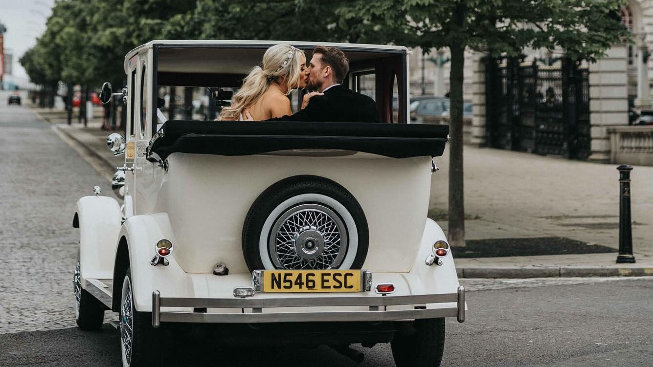 Rear view of Imperial car with spare wheel mounted at the rear of the vehicle, black soft top rear roof lowered showing Bride and Groom kissing inside the vehicle